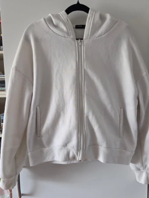 Vit hoodie med dragkedja - Säljer en enkel vit hoodie med dragkedja framtill och två fickor. Hoodien har ribbade muddar vid ärmslut och nederkant samt en stor huva. Perfekt för en chill och avslappnad stil. Materialet känns mjukt och bekvämt.