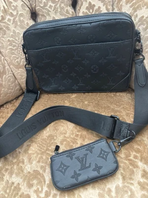 Svart crossbody-väska - Snygg svart crossbody-väska  i präglat läder med klassiskt monogrammönster. Väskan har en bred axelrem med logotyp och en matchande liten pouch som kan fästas på remmen. Slitage vid dragkedja osv vilket är vanligt då det är ej svart rakt igenom ingen av dem är . 