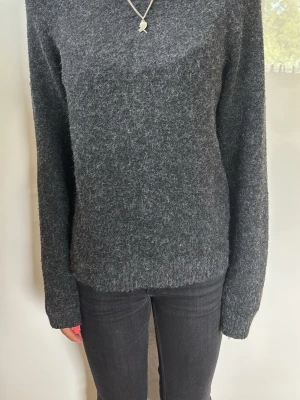 Mörkgrå stickad tröja - Mysig mörkgrå stickad tröja med lång ärm och enkel, rak passform. Tröjan har en mjuk och fluffig yta som ger en avslappnad look. Perfekt att matcha med jeans för en chill stil.
