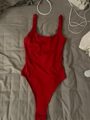 Röd body med djup rygg - Snygg röd body med breda axelband och hög skärning vid benen. Djup urringning i ryggen ger en trendig look. Tillverkad i stretchigt material som sitter bekvämt och formar sig efter kroppen. 