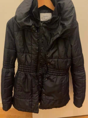 Svart pufferjacka från ONLY - Snygg svart pufferjacka från ONLY med hög krage och dragkedja framtill. Jackan har markerad midja med resår, quiltad design och breda muddar vid ärmslut. Perfekt för kalla dagar och har en modern, figurnära passform.
