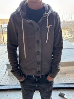 Mörkgrå hoodie från Hollister - Snygg mörkgrå hoodie från Hollister med knappar framtill och vit logga på bröstet. Stor huva med vita dragsnören och tryckt HOLLISTER-text längs ena ärmen. Tillverkad i mjukt bomullsmaterial, perfekt för en avslappnad stil. Storlek m men har krympt till xs