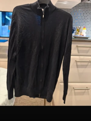 Svart ziptröja från Mersino - Svart ziptröja från Mersino med hög krage och långa ärmar. Tröjan är tunn och har en enkel design utan mönster, perfekt att slänga på sig när det är lite kyligt. Materialet är mjukt och känns bekvämt mot huden.
