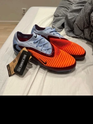 Nike fotbollsskor orange/blå - Säljer ett par grymma fotbollsskor från Nike med Dynamic Fit. Skorna har en cool design med orange och röda detaljer på ovandelen, ljusblå snörning och häl samt svarta Nike-loggor. Materialet är syntetiskt och anpassat för optimal passform och grepp på planen.