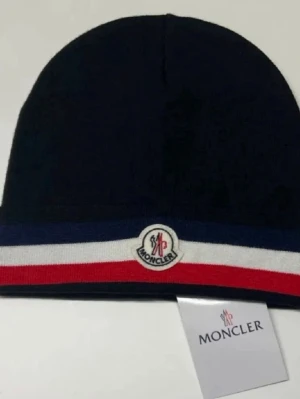 Mörkblå Moncler mössa med ränder - Snygg mörkblå mössa från Moncler med klassiska ränder i blått, vitt och rött längst ner. Mössan har Moncler-loggan framtill och är stickad i mjukt material som håller dig varm. Perfekt accessoar för dig som vill ha en stilren och trendig look.