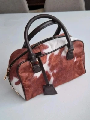 Handväska med konstgjord koskinn  - Unik handväska i brun- och vitmönstrat koskinn med svarta läderdetaljer och guldfärgade metalldelar. Väskan har två handtag, dragkedja upptill och förstärkt botten i svart läder med metallfötter. Avtagbart axelrem. Innerficka med dragkedja. Mått längd ca 22 cm Djup 10 cm Slutsåld på hemsidan  
