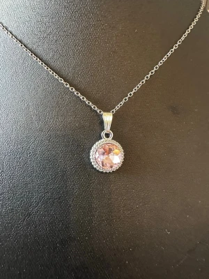 Silverhalsband med rosa sten - Snyggt halsband i silverfärgad metall med en rund, glittrande rosa sten som hänge. Hänget är dekorerat med små klara stenar runt den rosa stenen för extra bling. Perfekt för dig som gillar stilrena men ändå iögonfallande smycken.
