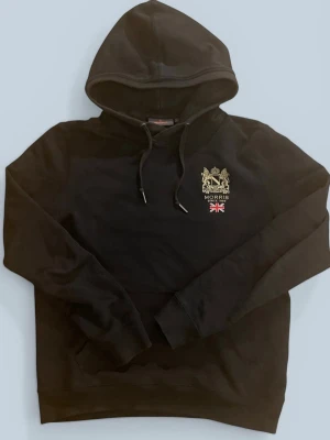 Svart hoodie från Morris - Svart hoodie från Morris med broderad logga och brittisk flagga på bröstet. Klassisk huva med dragsko och känguruficka framtill. Perfekt för en stilren och avslappnad look.