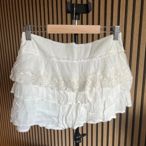 Vit volangskort från pull&bear - Supersöta vita skorts med flera lager volanger och fina spetsdetaljer framtill. Skortsen har en luftig och flowy känsla, perfekta för sommaren. Tillverkade i ett lätt tyg som ger en romantisk och trendig look.