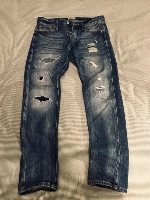 Blå slitna jeans med hål - Säljer ett par blå jeans med tydliga slitningar och flera hål på framsidan. Jeansen har klassisk femficksdesign, raka ben och en cool, tvättad look. Perfekta för dig som gillar en avslappnad och trendig streetstil.