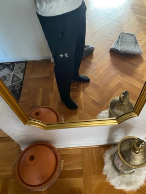 Svarta mjukisbyxor från Under Armour - Säljer ett par svarta mjukisbyxor från Under Armour med dragsko i midjan och två sidofickor. Byxorna har en avslappnad passform och den klassiska Under Armour-loggan i vitt på ena benet. Perfekta för chill eller träning.