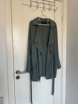 Grön trenchcoat  - Stilren grön  trenchcoat från bil bok med avslappnad passform, bred krage och knytskärp i midjan. Jackan har stora fickor och ett mjukt, tunt tyg som ger ett snyggt fall. Perfekt för lager på lager och trendiga outfits.