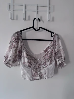 Blommig croppad blus med puffärm - Supersöt croppad blus med blommigt mönster i ljusa färger och puffiga kortärmar. Toppen har en feminin v-ringning och markerad midja med lätt korsettkänsla.