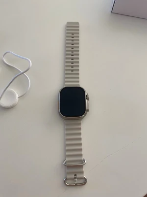 Apple Watch - Apple Watch Ultra med fyrkantig titanboett och ljusgrått silikonband. Klockan har en stor, tydlig skärm och en orange detalj på kronan. Perfekt för dig som vill ha en robust och snygg smartklocka med många funktioner. Box och sladdar medföljer. Priset går att diskutera vid snabb affär 