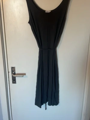 Svart långklänning med knytband - En enkel och stilren svart långklänning med rund halsringning och knytband i midjan. Klänningen är ärmlös och har ett mjukt, följsamt tyg som ger en avslappnad men snygg siluett. Perfekt för varma dagar eller när du vill känna dig bekväm men ändå chic.