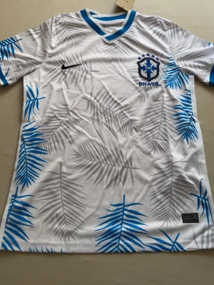 Vit Brasil fotbollströja Nike - Snygg vit fotbollströja från Nike med Brasil-logga på bröstet. Tröjan har blå och grå palmbladsmönster över hela, blå detaljer vid hals och ärmslut samt svart Nike-logga. Perfekt för dig som vill sticka ut på planen eller supporta Brasilien.