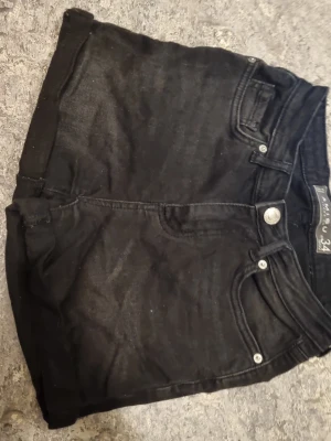 Svarta denimshorts  - Säljer ett par svarta denimshorts. Klassisk femficksmodell med knapp och dragkedja framtill. Shortsen har upprullade benslut och är perfekta till sommaren. Passar dig som gillar en enkel och stilren look.