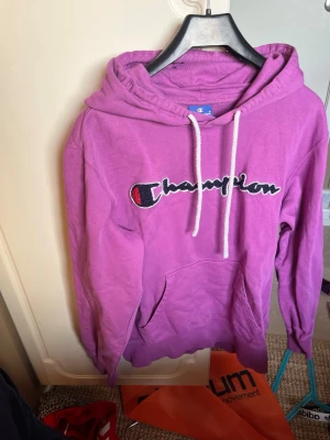 Lila Champion hoodie med huva - Säljer en lila hoodie från Champion med stor broderad logga på bröstet. Hoodien har vit dragsko i huvan och en klassisk känguruficka framtill. Tillverkad i mjukt bomullsmaterial och har en avslappnad passform. Perfekt för en chill och sportig look.