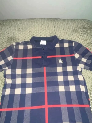 Rutig kortärmad t-shirt Burberry - Säljer en klassisk rutig t-shirt från Burberry med beige, mörkblå och röda detaljer. T-shirten har korta ärmar, krage och knappar framtill samt Burberry-logga på bröstet. Perfekt för dig som gillar stilrena och tidlösa plagg.