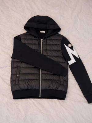 Svart Moncler cardiganjacka med huva - Cool svart cardiganjacka från Moncler med quiltad front, huva och dragkedja. Ärmarna är i mjukt tyg och ena ärmen har en vit zigzag-design samt Moncler-logga. Perfekt för dig som gillar streetstyle och vill ha något unikt.