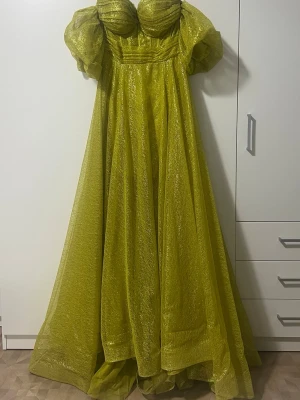 Glittrig gul långklänning med puffärm - Säljer en maxad gul långklänning med glittrigt tyg och stora puffärmar. Klänningen har en markerad byst, offshoulder-design och ett svepande fall. Perfekt för dig som vill sticka ut med en unik look.