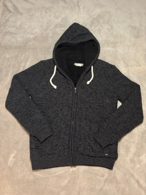 Stickad cardigan - Säljer en mörkgrå stickad cardigan med dragkedja och vit snörning i huvan. Stickad struktur på både ärmar och framsida ger ett coolt mönster. Perfekt för dig som gillar en avslappnad och trendig look.