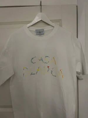 Vit t-shirt från Casablanca - Vit t-shirt från Casablanca med färgglad, blommig text på bröstet. Klassisk passform och rund halsringning. Perfekt för en stilren och trendig look.