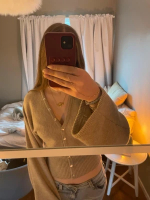 Beige kofta med vida ärmar - Säljer en beige kofta med v-ringning och vida ärmar. Koftan har ribbstickad nederkant och knäpps med pärlemorskimrande knappar framtill. Perfekt att styla med jeans för en chill look. Säljer för att den är lite för stor och inte kommer till användning. Väldigt bra skick, bara använd ett fåtal gånger.