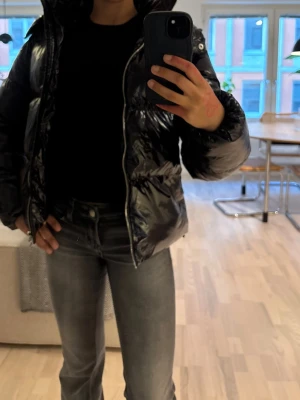 Svart glansig pufferjacka med huva(Nelly) - Säljer en svart, glansig pufferjacka med huva och dragkedja framtill. Jackan har en boxig passform och breda kanaler som ger en riktigt puffig look. Perfekt för kalla dagar när du vill hålla värmen och ändå se stylish ut. Knappt använd och köptes för 799💕