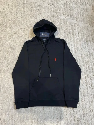 Svart hoodie från Polo Ralph Lauren - Svart hoodie från Polo Ralph Lauren med klassisk huva och dragsnören. Har en röd broderad logga på bröstet och en stor magficka framtill. Tillverkad i mjukt bomullsmaterial och har ribbade muddar vid ärmslut och nederkant.