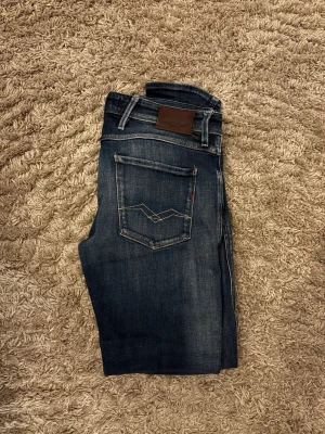 Mörkblå  jeans från Replay - Säljer ett par mörkblå  jeans från Replay med snygga slitningar och kontrastsömmar. Jeansen har klassisk femficksmodell, dragkedja och knapp i midjan samt Replay-lapp bak i läderimitation. Perfekta för en trendig och avslappnad look.