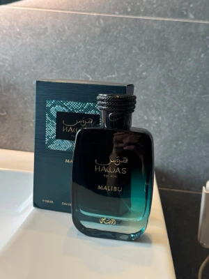 Hawas For Him Malibu parfym - Snygg parfymflaska från Hawas, modellen Malibu. Flaskan är rektangulär med rundade kanter och har en snygg blå-turkos färg som tonar ut mot svart upptill. Märkets logga och namn står i guld på flaskan. Kommer i matchande förpackning.