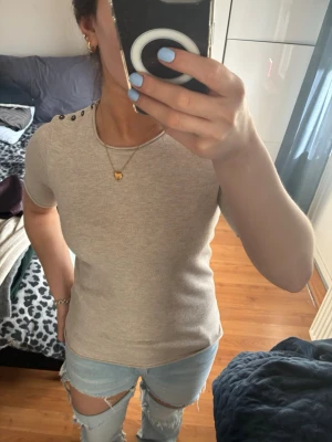 Beige stickad tröja med knappar H&M - Beige stickad tröja från H&M Basics i storlek S. Rund halsringning och korta ärmar. Snygga mörka knappar på ena axeln ger en extra detalj. Perfekt för dig som gillar enkel och stilren design med en twist.