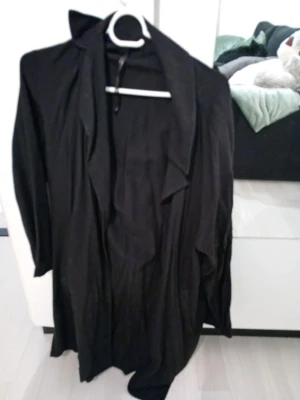 Svart lång trenchcoat med lös passform - Säljer en svart lång trenchcoat med avslappnad och lös passform. Rocken har bred krage och faller snyggt framtill. Perfekt för dig som gillar en stilren och minimalistisk look.