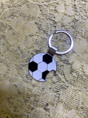 Nyckelring med flasköppnare i fotbollsmotiv - Säljer en unik nyckelring i metall med ett fotbollsmotiv i svart och vitt. Nyckelringen har en rund form och en detalj som ser ut som ett bett i kanten, vilket ger den en rolig twist. Perfekt för dig som älskar fotboll och vill ha något personligt till dina nycklar.