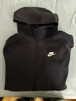 Blått Nike zip hoodie med huva - Säljer en svart hoodie från Nike med dragkedja framtill och klassisk vit Nike-logga på bröstet. Hoodien har huva och två fickor framtill. Tillverkad i mjukt och bekvämt material, perfekt för en avslappnad streetwear-look.