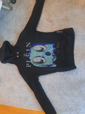 Svart hoodie Philipp Plein blå döskalle - Säljer en svart hoodie från Philipp Plein med en stor blå och silverglittrig döskalle på bröstet. Hoodien har dragsko i huvan och coola detaljer med text och mönster framtill. Perfekt för dig som gillar streetwear och vill sticka ut.