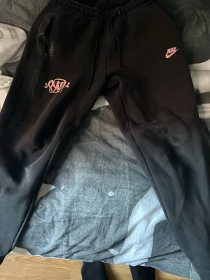 Svarta Nike mjukisbyxor med rosa tryck - Säljer ett par svarta mjukisbyxor från Nike med rosa logga och tryck på vänster ben. Byxorna har dragsko i midjan, fickor med dragkedja och är tillverkade i mjukt material. Perfekta för chill eller streetwear-stil.