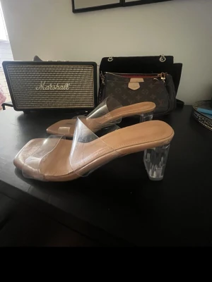 Transparenta sandaler med klack - Säljer ett par trendiga sandaler med transparent rem och klar blockklack. Sulan är beige och har en fyrkantig tå för en modern look. Perfekta för dig som vill ha en clean och stilren vibe till outfiten.