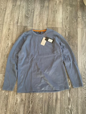 Blå långärmad tröja från BOSS - Snygg blå långärmad tröja från BOSS med rund halsringning och diskret logga på bröstet. Tröjan har en enkel och stilren design, perfekt för en avslappnad look. Tillverkad i mjukt material för extra komfort. Tröjan är i storlek XXL, men sitter som XL