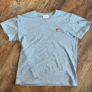 Grå t-shirt från Ami Paris - Snygg grå t-shirt från Ami Paris med klassisk passform och liten röd broderad logga på bröstet. Tillverkad i mjuk bomull och har rund hals samt korta ärmar. Perfekt basplagg för en clean och stilren look.