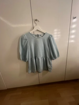 Ljusblå blus med puffärm - Supersöt ljusblå blus med puffiga kortärmar och rund hals. Blusen har en lös passform och volang nedtill samt knytning i nacken. Tillverkad i ett mjukt tyg som känns skönt mot huden. Perfekt att styla med jeans eller kjol.