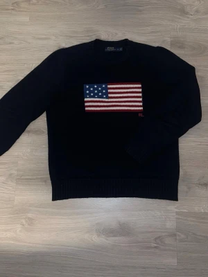 Mörkblå stickad tröja Polo Ralph Lauren - Snygg mörkblå stickad tröja från Polo Ralph Lauren med amerikansk flagga i rött, vitt och blått på bröstet. Tröjan har rund halsringning, ribbade muddar och RL-broderi. Perfekt för dig som gillar klassisk preppy stil.