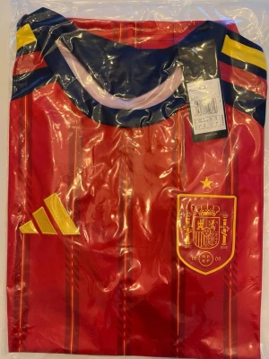 Spaniens matchtröja 2026. Helt ny! - Helt ny! Säljer en röd matchtröja från Adidas med Spaniens landslagsmärke på bröstet och gula detaljer. Tröjan har mörkblå och gula inslag vid axlarna samt Adidas logga i gult. Perfekt för fotbollsfans som vill stötta Spanien.
