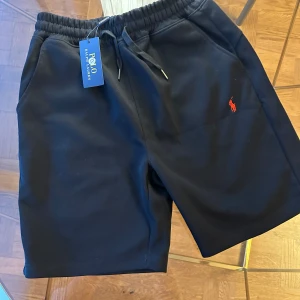 Svarta shorts från Polo Ralph Lauren - Svarta shorts från Polo Ralph Lauren med elastisk midja och snörning. Klassisk röd broderad logga på vänster ben. Mjukt material och två sidofickor. Perfekta för en avslappnad stil.