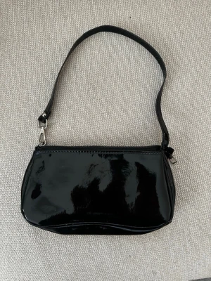 Svart lackad axelväska - Snygg svart handväska i glansigt lackmaterial med enkel axelrem och silverfärgade metalldetaljer. Väskan har en klassisk rektangulär form och stängs med dragkedja upptill. Perfekt för dig som gillar en stilren och trendig look.