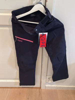 Mörkblå täckbyxor från EQPE - Snygga mörkblå täckbyxor från EQPE med en rosa dragkedja och logga på vänster ben. Byxorna har rak passform och är tillverkade i ett slitstarkt, vattenavvisande material. Perfekta för kalla dagar utomhus och vinteraktiviteter.