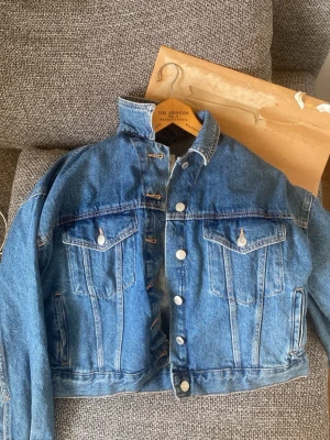 Blå klassisk jeansjacka - Snygg blå jeansjacka med raka axlar och två stora bröstfickor med lock och knapp. Jackan har silverfärgade knappar framtill och vid fickorna, samt en hög krage för extra attityd. Perfekt för dig som vill ha en tidlös och cool look.