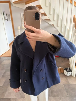 Mörkblå dubbelknäppt kappa - Säljer en mörkblå kappa i storlek XS/S med dubbelknäppning och bred krage. Kappan har stora knappar framtill, två snedställda fickor och uppvikta ärmslut. Modellen är boxig och lite kortare, vilket ger en modern och avslappnad look. Materialet känns som ullblandning.