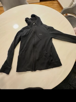 Svart hoodie med dragkedja från BOSS - Snygg svart hoodie från BOSS med dragkedja framtill och två fickor. Klassisk design med huva och justerbara snören. Diskret BOSS-logga på bröstet. Perfekt för en avslappnad och stilren look.